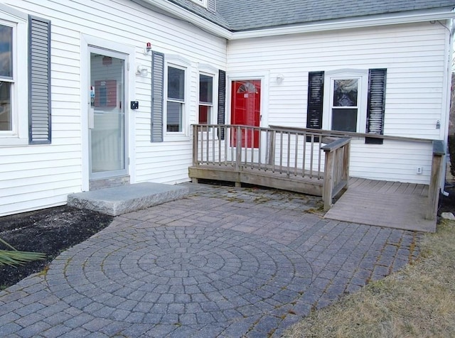 view of exterior entry with a deck, a patio, and roof with shingles