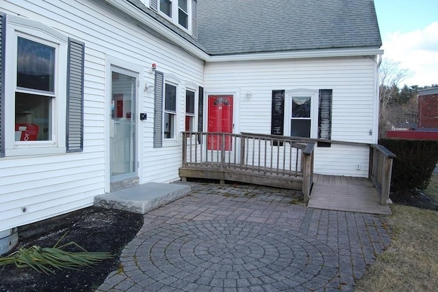 view of exterior entry featuring a deck, a shingled roof, and a patio area