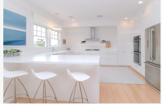 kitchen with a peninsula, light countertops, wall chimney exhaust hood, white cabinetry, and appliances with stainless steel finishes