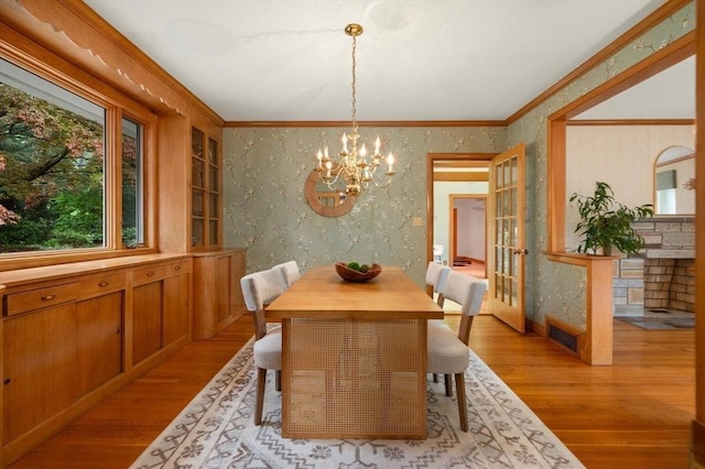 dining space featuring a chandelier, light wood-style flooring, crown molding, wallpapered walls, and french doors
