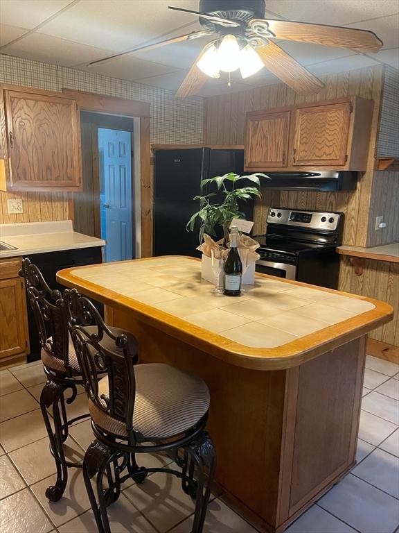 kitchen featuring light tile patterned floors, black fridge, stainless steel range with electric cooktop, ceiling fan, and wood walls