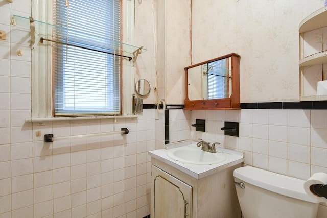 half bathroom featuring tile walls, wallpapered walls, toilet, wainscoting, and vanity