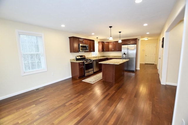 kitchen with pendant lighting, a center island, dark wood-type flooring, light stone countertops, and appliances with stainless steel finishes