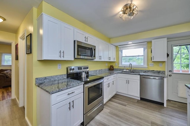 kitchen featuring appliances with stainless steel finishes, a sink, white cabinets, light wood-style floors, and dark stone counters