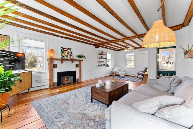 living room with ceiling fan, beam ceiling, baseboard heating, a glass covered fireplace, and wood-type flooring