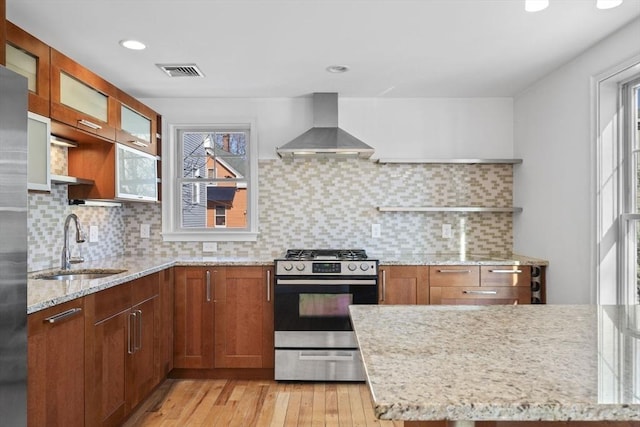 kitchen with decorative backsplash, stainless steel gas stove, wall chimney range hood, light stone counters, and sink