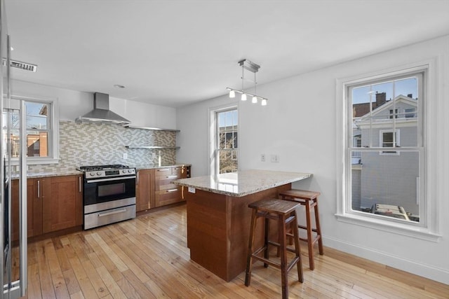 kitchen with a kitchen bar, backsplash, stainless steel gas stove, wall chimney range hood, and light stone counters