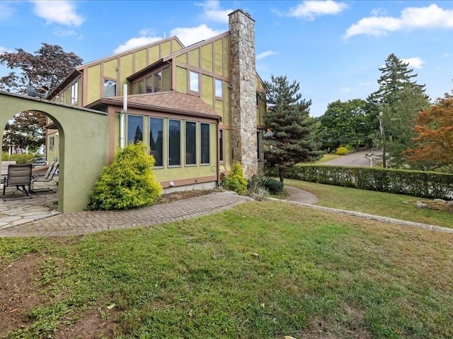 rear view of property featuring a shingled roof, a lawn, a chimney, a patio area, and stucco siding