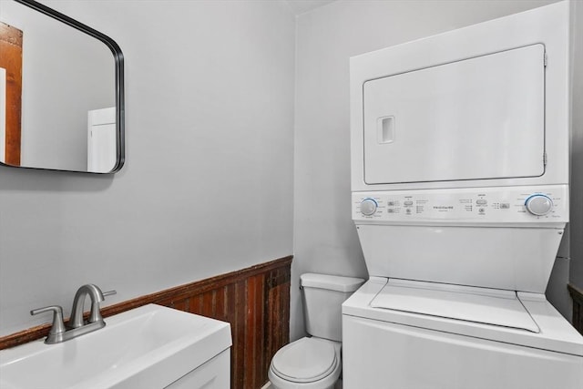 bathroom with stacked washer / dryer, toilet, a sink, and a wainscoted wall
