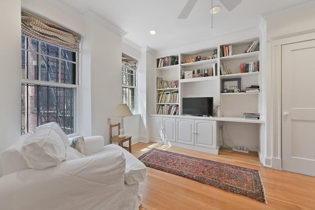 living area with crown molding, light wood-style floors, a ceiling fan, built in shelves, and baseboards