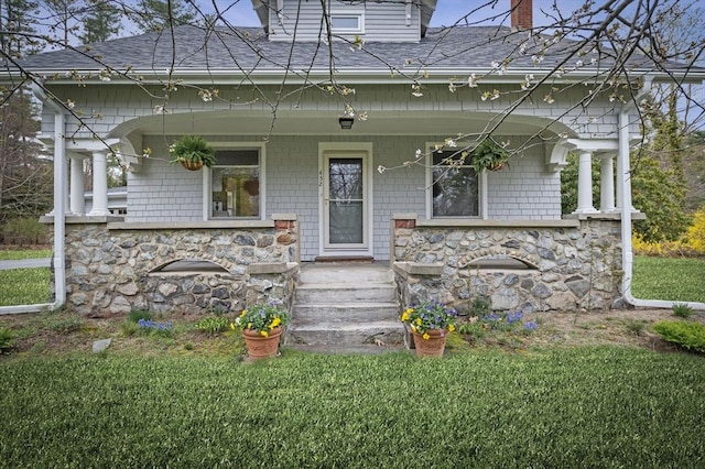 view of front facade with covered porch, a front yard, stone siding, roof with shingles, and a chimney
