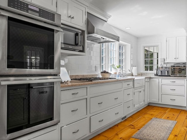 kitchen featuring light wood-type flooring, wall chimney exhaust hood, white cabinets, a sink, and stainless steel appliances