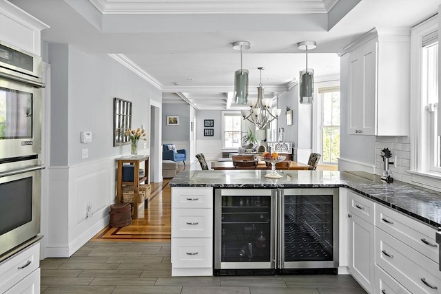 kitchen with beverage cooler, a raised ceiling, a peninsula, white cabinetry, and a wainscoted wall