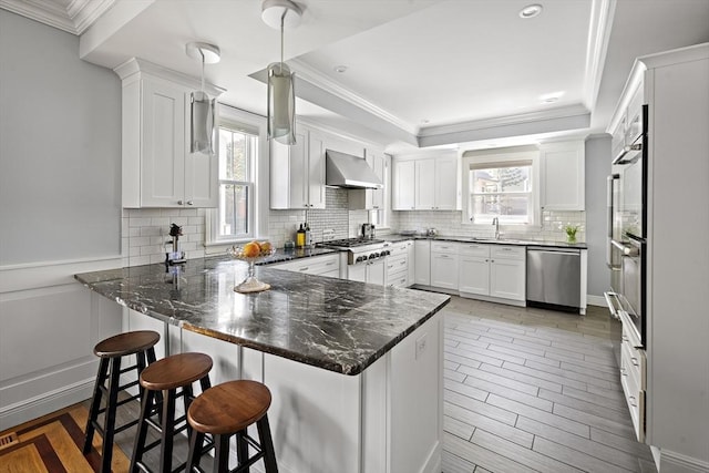 kitchen featuring appliances with stainless steel finishes, a peninsula, a sink, wall chimney exhaust hood, and healthy amount of natural light