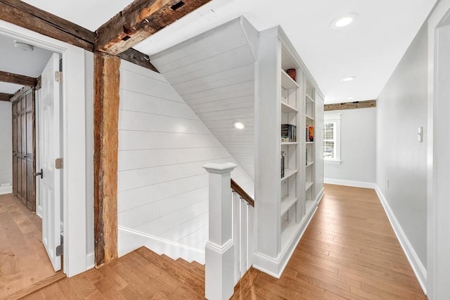 hall featuring light wood-style flooring, baseboards, beamed ceiling, and an upstairs landing