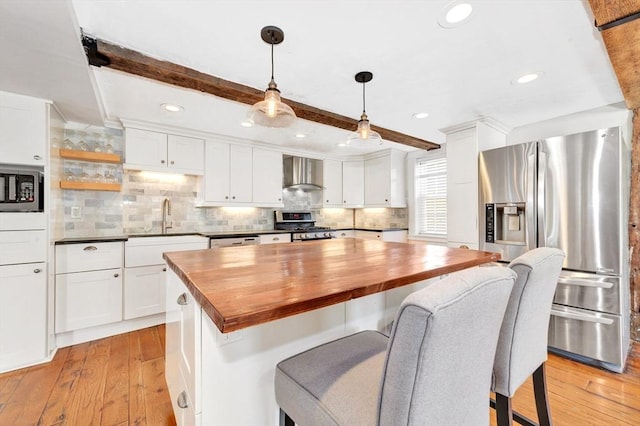 kitchen featuring butcher block counters, appliances with stainless steel finishes, light wood-type flooring, beam ceiling, and wall chimney exhaust hood