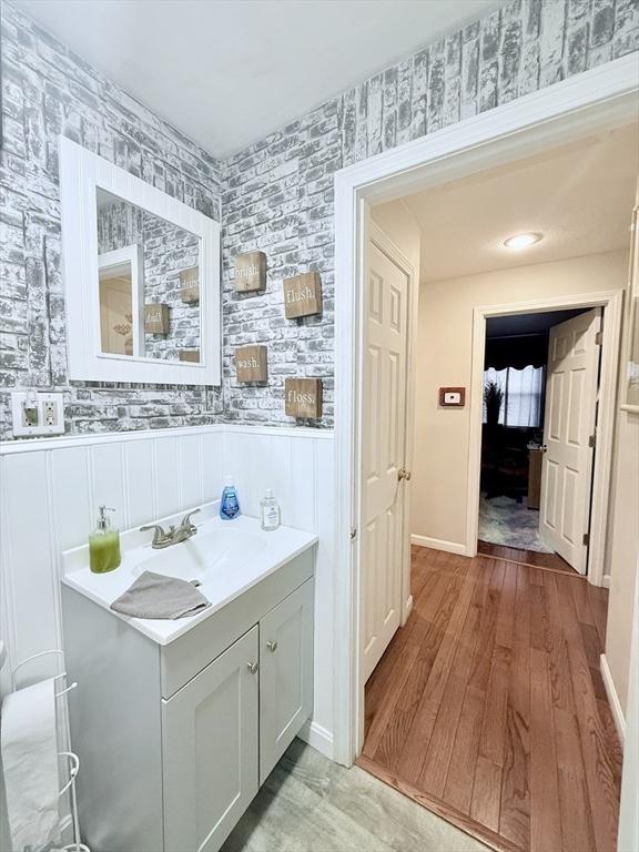 bathroom with wallpapered walls, vanity, wood finished floors, and wainscoting