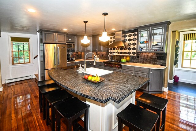 kitchen with built in appliances, dark hardwood / wood-style floors, a kitchen island with sink, baseboard heating, and wall chimney range hood