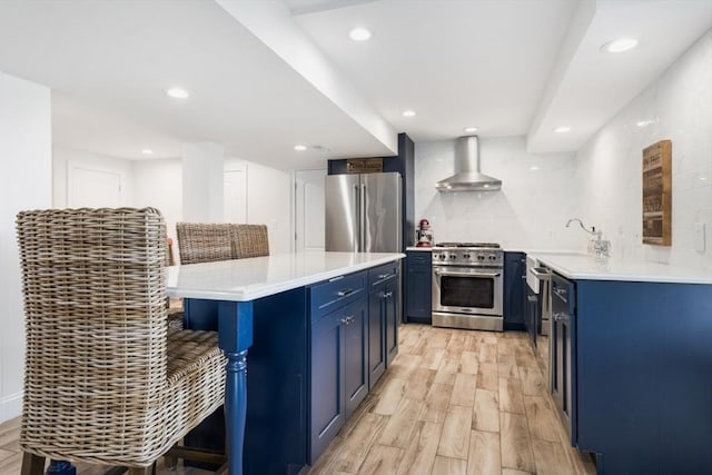 kitchen with blue cabinetry, appliances with stainless steel finishes, a kitchen bar, and wall chimney range hood