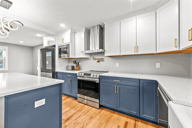 kitchen with white cabinets, wall chimney range hood, blue cabinets, appliances with stainless steel finishes, and light hardwood / wood-style floors