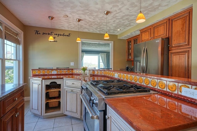 kitchen with brown cabinets, hanging light fixtures, appliances with stainless steel finishes, light tile patterned floors, and glass insert cabinets