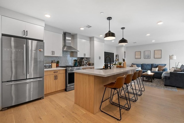 kitchen featuring pendant lighting, a kitchen island with sink, wall chimney exhaust hood, white cabinetry, and stainless steel appliances