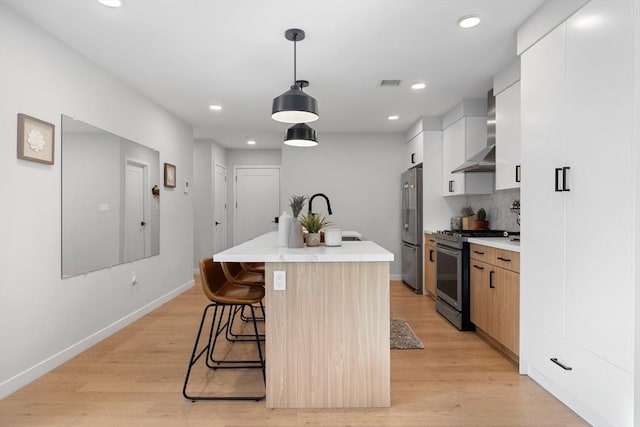 kitchen featuring appliances with stainless steel finishes, wall chimney exhaust hood, white cabinetry, hanging light fixtures, and an island with sink