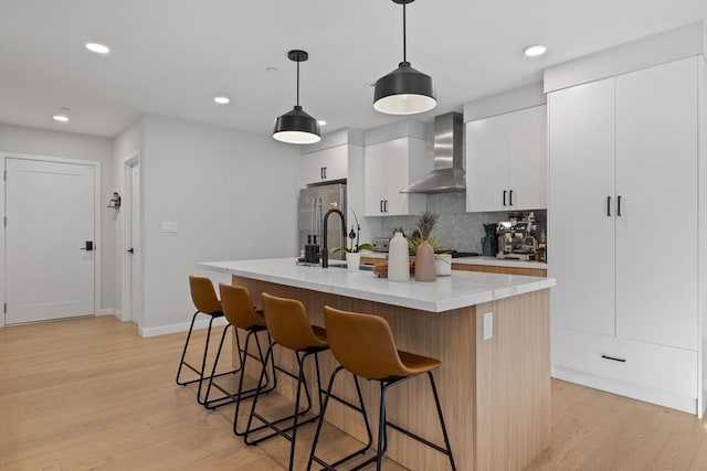 kitchen featuring wall chimney range hood, hanging light fixtures, stainless steel fridge, an island with sink, and white cabinetry