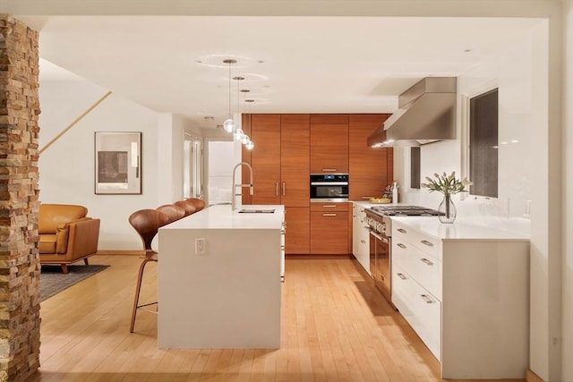 kitchen featuring a sink, stainless steel appliances, wall chimney range hood, brown cabinets, and light wood-style flooring