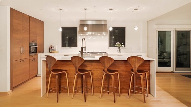 bar area with wall chimney range hood, oven, light wood finished floors, pendant lighting, and a sink