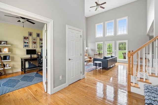 entryway featuring a ceiling fan, light wood-type flooring, a towering ceiling, stairs, and baseboards