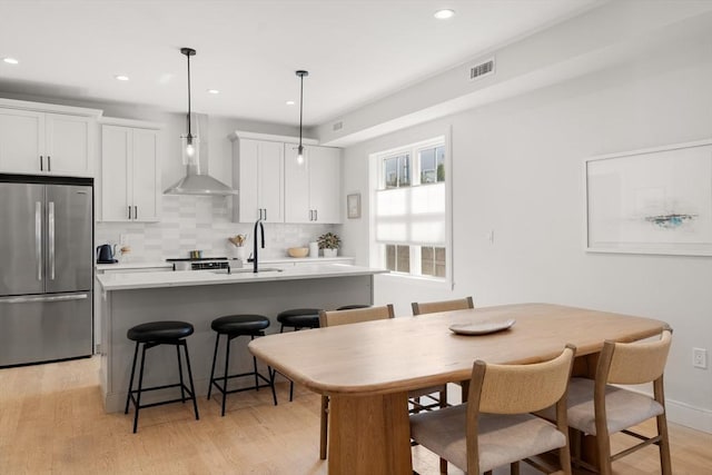 kitchen featuring stainless steel fridge, wall chimney range hood, an island with sink, a sink, and decorative backsplash