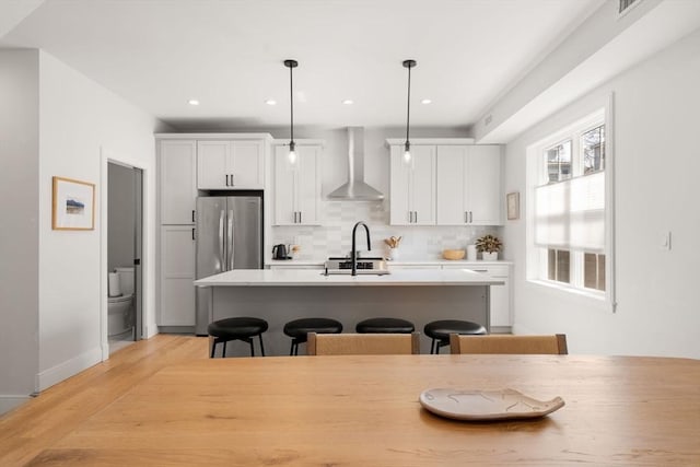 kitchen with wall chimney exhaust hood, freestanding refrigerator, a kitchen breakfast bar, a center island with sink, and light wood-type flooring