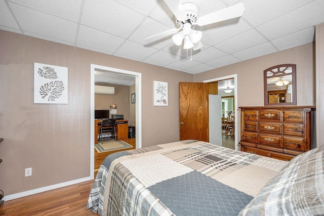 bedroom featuring a wall mounted AC, a drop ceiling, wood finished floors, baseboards, and ceiling fan