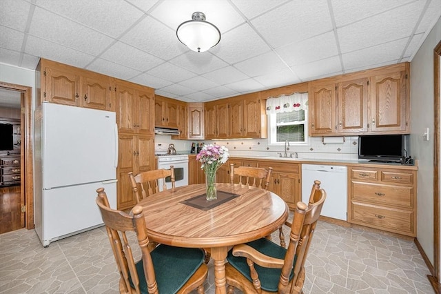 kitchen with white appliances, a sink, under cabinet range hood, a paneled ceiling, and stone flooring
