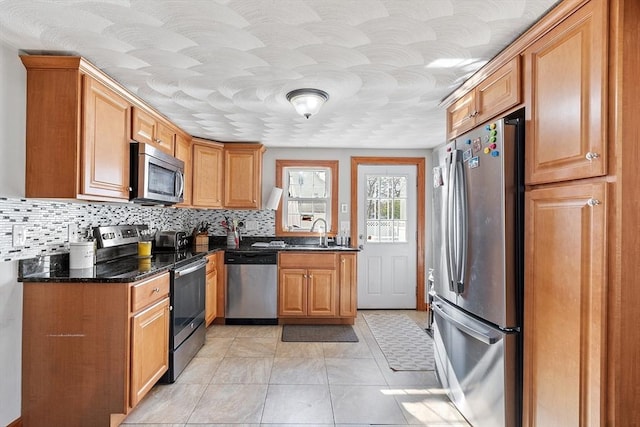 kitchen featuring dark stone counters, a sink, brown cabinets, backsplash, and appliances with stainless steel finishes