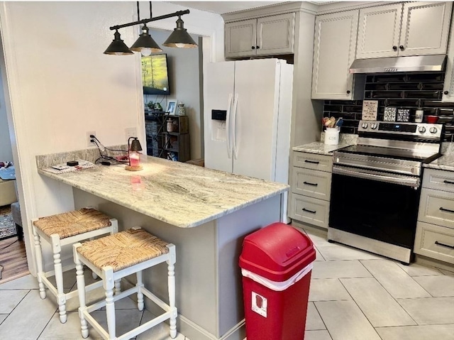 kitchen with light tile patterned floors, white fridge with ice dispenser, stainless steel electric stove, under cabinet range hood, and a kitchen bar