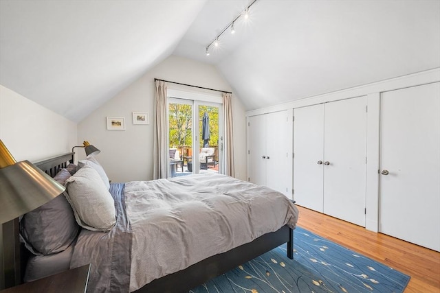 bedroom with access to exterior, vaulted ceiling, rail lighting, wood finished floors, and two closets