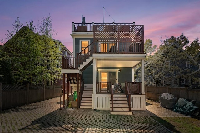 back of house at dusk featuring fence private yard, a balcony, stairway, and covered porch