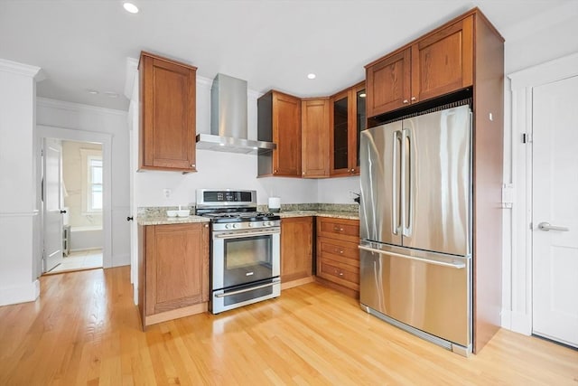 kitchen with appliances with stainless steel finishes, wall chimney range hood, light wood-style flooring, brown cabinetry, and glass insert cabinets