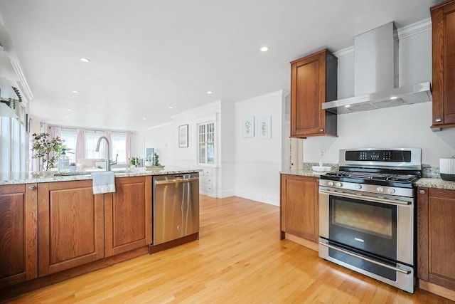 kitchen featuring stainless steel appliances, wall chimney exhaust hood, light wood-style flooring, brown cabinetry, and ornamental molding