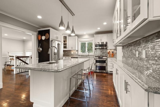 kitchen with white cabinets, an island with sink, glass insert cabinets, light stone countertops, and stainless steel appliances