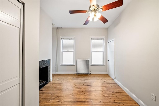 spare room featuring radiator, a fireplace, light wood finished floors, a ceiling fan, and baseboards