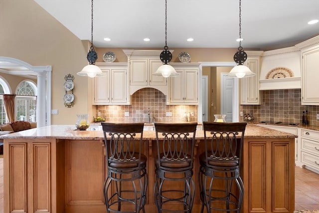 kitchen featuring light stone counters, cream cabinets, a kitchen island with sink, black electric cooktop, and hanging light fixtures