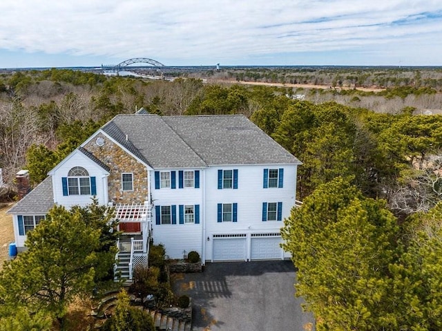 view of front of home featuring driveway, an attached garage, a shingled roof, stone siding, and a view of trees