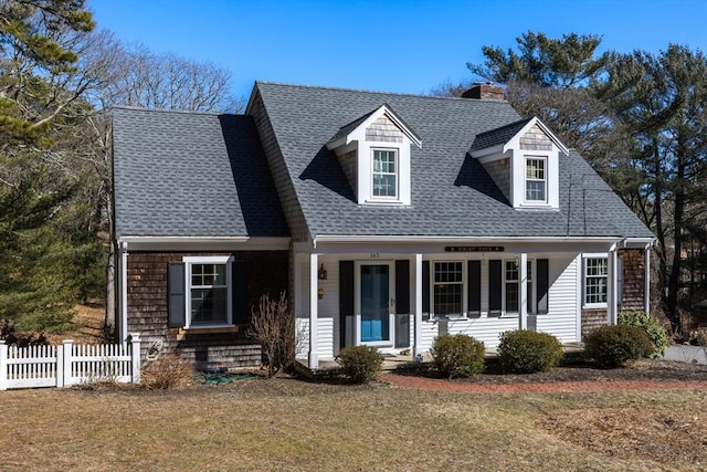 new england style home featuring a front lawn, fence, covered porch, a shingled roof, and a chimney