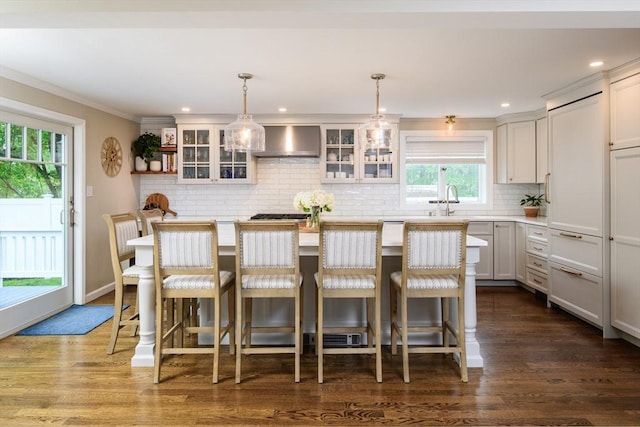kitchen featuring wall chimney exhaust hood, backsplash, light countertops, glass insert cabinets, and ornamental molding