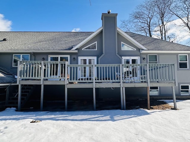 snow covered back of property featuring french doors, roof with shingles, a chimney, a wooden deck, and stairs