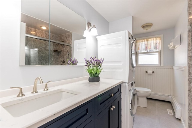 bathroom with tile patterned flooring, a wainscoted wall, toilet, stacked washer and clothes dryer, and a stall shower