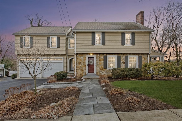 colonial inspired home featuring an attached garage, a chimney, stone siding, aphalt driveway, and a lawn